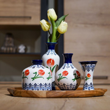 Vier verschillende vaasjes. Een witte achtergrond met onderaan en bovenaan Delfts blauw versiering. In het midden van de vaas rondom oranje tulpen. Een kelkvaasje, buikvaasje, rond vaasje en een slank vaasje.